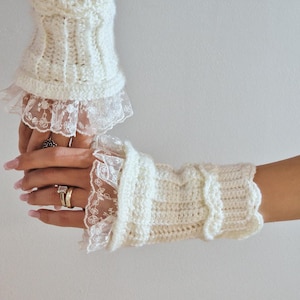 Könnte beinhalten: Cremefarbene, gehäkelte Fingerhandschuhe mit zarter Spitzenborte. Die Handschuhe haben ein Rippenmuster am oberen Teil und einen gewellten Rand. Die Spitze verleiht dem handgefertigten Accessoire einen Hauch von Eleganz.