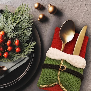 May include: A Christmas table setting featuring a red and green stocking-shaped cutlery holder, gold utensils, and gold baubles. A black plate with greenery and red berries enhances the festive atmosphere.