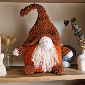 Op de afbeelding: Een handgemaakte pluche kabouterpop met een bruine hoed en armen, een oranje lichaam en een witte baard. De kabouter heeft een witte pomponneus en bruine voeten. De pop staat op een houten plank.