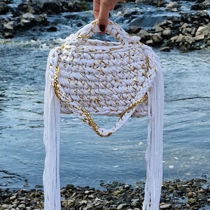 Peut inclure: Un sac à main crocheté blanc et doré, de forme arrondie et avec une poignée. Le sac présente des accents dorés et de longues franges blanches. Le sac est présenté devant une rivière.