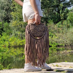 May include: A handmade, fringed handbag in a neutral taupe color. The bag features a woven top with a rounded shape and a handle. Long, soft fringe hangs from the bottom, creating a bohemian style. The bag is carried outdoors.