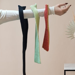 Op de afbeelding: Drie gebreide haarbanden in zwart, lichtgroen en rood-gele gradiëntkleuren. De haarbanden worden gepresenteerd op een wit oppervlak tegen een lichtroze achtergrond. De haarbanden zijn gemaakt van een getextureerd gebreid materiaal.