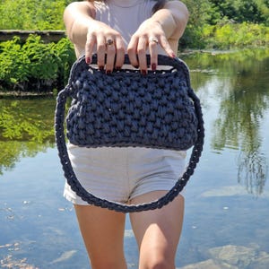 May include: A dark grey crocheted handbag with a rectangular shape and a woven texture. The bag has a top handle and a long shoulder strap. The background shows a body of water and greenery.