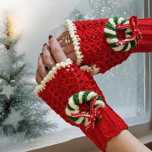 May include: Red crochet fingerless gloves with a cream-colored scalloped trim. Each glove features a small wreath with green and white stripes and a red bow. The gloves are designed for warmth and style.