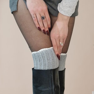 May include: A pair of light gray crochet leg warmers with a white trim, worn over black tights and black boots. The leg warmers are visible from the knees down.
