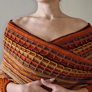 May include: A handmade crochet shawl in warm autumn colors. The shawl features a geometric pattern in shades of orange, brown, and tan. The wrap is draped over the shoulders, showcasing the intricate stitch work and color variations. A cozy and stylish accessory.