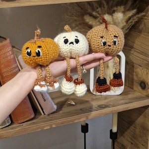May include: Three handmade crochet pumpkins in shades of orange, cream, and brown, each with a smiling face and dangling legs. The pumpkins are displayed on a wooden shelf, showcasing a whimsical autumn theme.