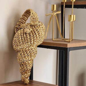 Op de afbeelding: Een gouden, geweven handtas in de vorm van een zeeschelp, met een handvat en een knoopsluiting. De tas is gemaakt van een glanzend, getextureerd materiaal. Een goudkleurige kandelaar staat op een houten plank.