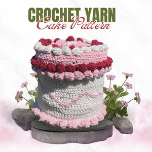 May include: A crocheted cake pattern with the text "CROCHET YARN Cake Pattern." The cake is white, pink, and red, with decorative trim and a scalloped edge. The cake sits on a gray stone base, with pink flowers in the background.