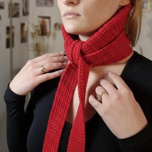 May include: A vibrant red knitted scarf tied around the neck. The scarf is made of a thick, textured yarn and drapes down the front. The person is wearing a black long-sleeve shirt and rings.
