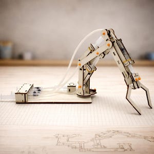 May include: A wooden robotic arm kit with a base, syringes, and tubing. The arm is made of light-colored wood with orange accents and a claw-like gripper. The kit is designed for educational or hobbyist use.