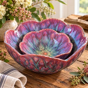 May include: Two nested ceramic bowls with a scalloped, flower-like rim. The glaze features a vibrant, swirling blend of deep magenta, purple, and teal colors with a glossy finish. The interior of the bowls displays a radiating pattern of colors.