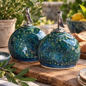 May include: Two round ceramic oil dispensers featuring a glossy, mottled glaze in shades of deep teal, blue, and green. Each dispenser is topped with a silver-colored metal pour spout with a hinged lever mechanism for controlled liquid dispensing.
