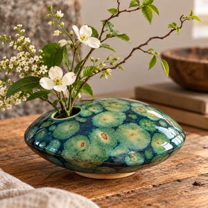 May include: A low, disc-shaped ceramic vase with a glossy, crystalline glaze featuring a pattern of teal and dark blue circular spots. The vase has a small, centered opening holding a branch with white flowers and green leaves.