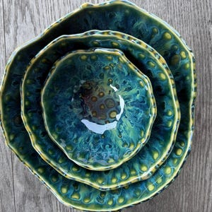 May include: A set of three nested ceramic bowls with an organic, wavy rim shape. The glaze features a deep teal and forest green color palette with a reactive, mottled pattern that creates a textured, bubble-like appearance across the surface.