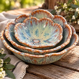 May include: A set of three nested ceramic bowls with a scalloped, flower-like edge. The bowls feature a reactive glaze with a blend of light blue, cream, and brown tones, creating a variegated finish that highlights the textured, wavy rims.
