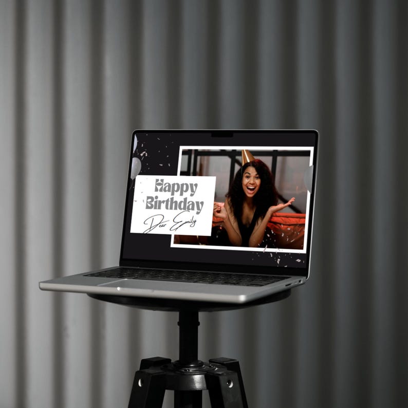May include: A silver laptop shows a birthday message reading "Happy Birthday Dear Emily." The screen features a smiling person wearing a party hat. The laptop is on a black tripod stand against a grey backdrop.
