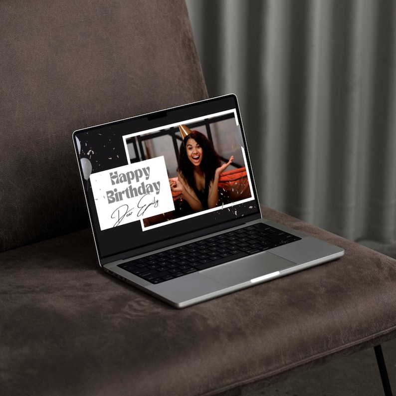 May include: A silver laptop showing a birthday message on its screen. The screen displays a photo of a person wearing a party hat, with the words "Happy Birthday Dear Emily". The laptop is on a brown upholstered surface.