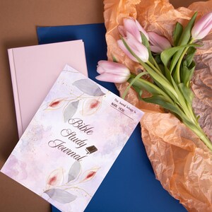 Könnte beinhalten: Ein Bibelstudien-Journal mit floralem Design und dem Text "Bible Study Journal". Das Cover ist mit Blättern im Aquarellstil und goldenen Akzenten verziert. Ein rosa Buch und ein blaues Buch sind ebenfalls im Bild, zusammen mit rosa Tulpen.