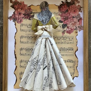 May include: Framed artwork featuring a paper sculpture of a woman in a dress made from sheet music. The figure has a yellow top, a blue scarf, and a bow. Floral accents and musical notes are visible.