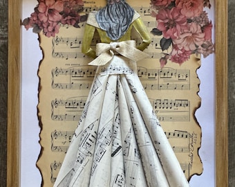 Può includere: Opera d'arte incorniciata con una scultura di carta di una donna in un vestito fatto di spartiti musicali. La figura ha un top giallo, una sciarpa blu e un fiocco. Sono visibili elementi floreali e note musicali.