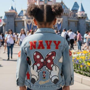 May include: A light blue denim jacket featuring the word NAVY in red block letters across the back. Below the text is a large, glittery red and white polka-dot bow held by two white gloved hands, with a small Minnie Mouse figure and star patches.