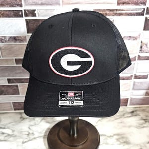 May include: Black trucker hat with a mesh back and a curved brim. The front features a white and red oval patch with a white "G" inside. The hat has an adjustable strap and a "Richardson" label.