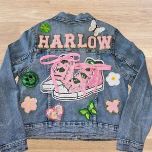 May include: A light blue denim jacket with patches and the name "HARLOW" in pink letters. The jacket features patches of a smiley face, flowers, butterflies, and pink sequined sneakers with pink laces.