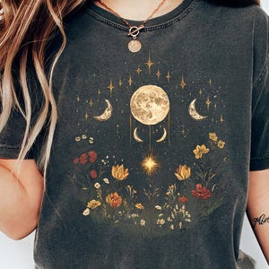Puede incluir: Camiseta gris oscuro con un diseño celestial. El gráfico incluye una luna llena, lunas crecientes, estrellas y flores silvestres en tonos amarillos, rojos y naranjas. La camiseta tiene un aspecto vintage y desgastado.