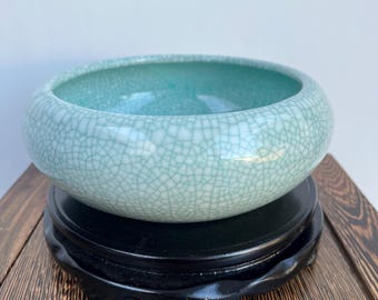 May include: A light blue ceramic bowl with a crackled glaze. The bowl is sitting on a black wooden stand.