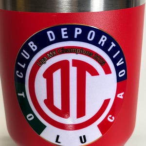 May include: A red insulated tumbler with a silver rim and lid. The tumbler features a circular emblem with the words "CLUB DEPORTIVO TOLUCA" and "Liga MX Champions 2025" in white text. The emblem has a red and white logo.