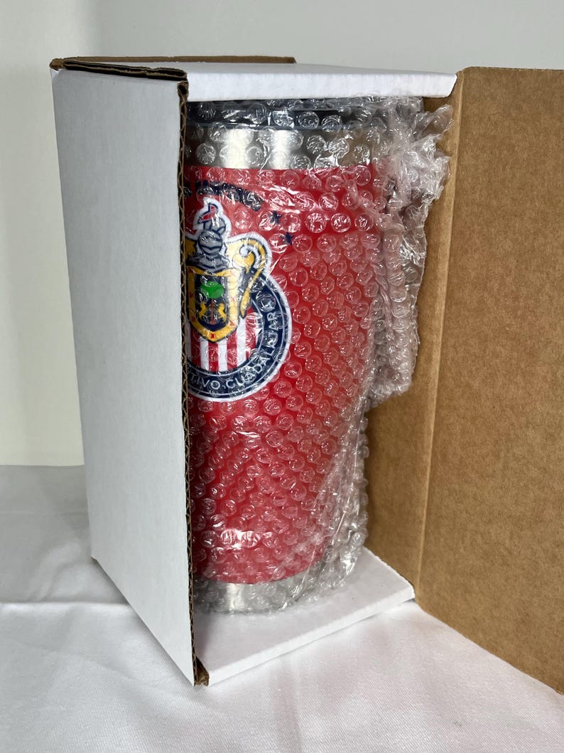Puede incluir: Un vaso t&eacute;rmico rojo con un logotipo de equipo, empaquetado en una caja de cart&oacute;n blanca. El vaso est&aacute; envuelto en pl&aacute;stico de burbujas para protecci&oacute;n. El logotipo del equipo es un escudo con un escudo y las palabras "Club Deportivo Guadalajara".