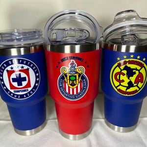 Puede incluir: Tres vasos aislados con logotipos de equipos. Uno es azul con un logotipo blanco y rojo, otro es rojo con un escudo del equipo y el tercero es azul con un logotipo amarillo y rojo. Cada uno tiene una tapa transparente y una base plateada.