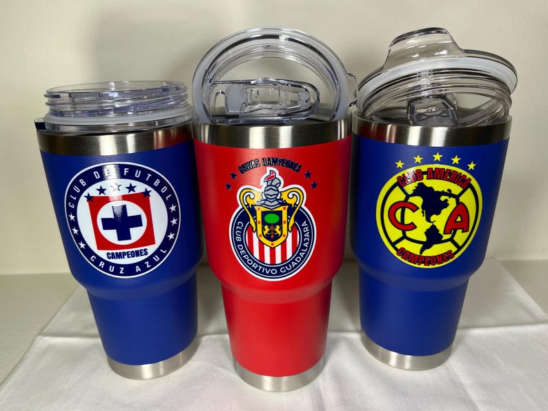 Puede incluir: Tres vasos aislados con logotipos de equipos. Uno es azul con un logotipo blanco y rojo, otro es rojo con un escudo del equipo y el tercero es azul con un logotipo amarillo y rojo. Cada uno tiene una tapa transparente y una base plateada.