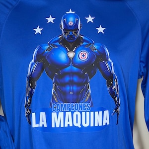 May include: Royal blue hoodie with a futuristic figure design, team logo, and white stars. The text "CAMPEONES LA MAQUINA" is displayed in white. The hoodie has long sleeves and a hood. The design is centered on the chest.