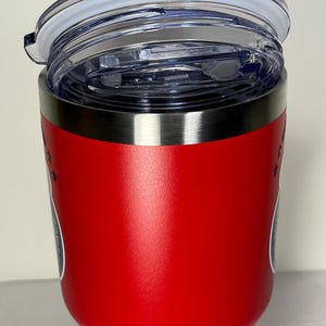 Puede incluir: Un vaso t&eacute;rmico rojo con borde plateado y tapa de pl&aacute;stico transparente. El vaso tiene un dise&ntilde;o con estrellas y un emblema circular. La tapa est&aacute; parcialmente abierta, mostrando el interior.