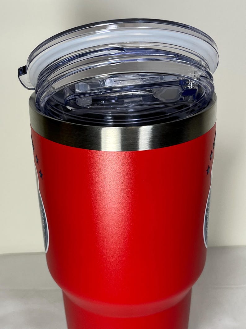 Puede incluir: Un vaso t&eacute;rmico rojo con borde plateado y tapa de pl&aacute;stico transparente. El vaso tiene un dise&ntilde;o con estrellas y un emblema circular. La tapa est&aacute; parcialmente abierta, mostrando el interior.