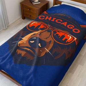 May include: A blue blanket with a bear wearing orange sunglasses, the word "CHICAGO" above. The bear is brown and tan, with the city skyline reflected in the sunglasses. The blanket is on a bed.