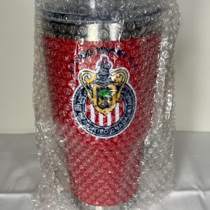 Puede incluir: Un vaso t&eacute;rmico rojo con tapa negra, envuelto en pl&aacute;stico de burbujas. El vaso presenta un logotipo del equipo con un escudo, un escudo y texto. El logotipo es blanco, dorado, azul y verde. El vaso est&aacute; dise&ntilde;ado para mantener las bebidas calientes o fr&iacute;as.