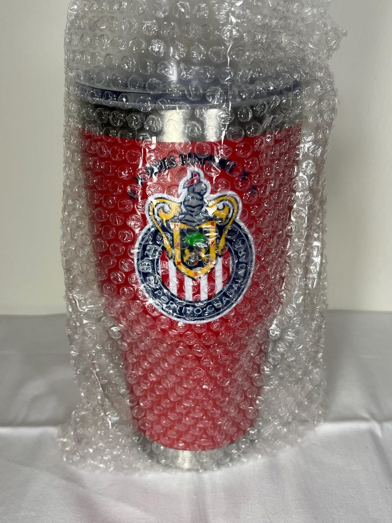 Puede incluir: Un vaso t&eacute;rmico rojo con tapa negra, envuelto en pl&aacute;stico de burbujas. El vaso presenta un logotipo del equipo con un escudo, un escudo y texto. El logotipo es blanco, dorado, azul y verde. El vaso est&aacute; dise&ntilde;ado para mantener las bebidas calientes o fr&iacute;as.