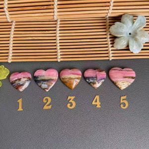 May include: Five polished heart-shaped stones arranged in a row, each featuring bands of pink, white, brown, and black. Below each stone is a gold-colored number from one to five. A light-colored carved stone flower sits in the upper right corner.
