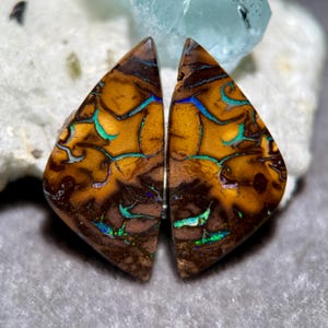 Peut inclure: Deux pierres d'opale triangulaires. Les opales présentent une couleur de base marron avec des motifs vibrants orange, bleu et vert. Les pierres sont taillées et polies, révélant leurs structures internes uniques. Les opales sont posées sur un fond neutre.