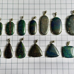 Op de afbeelding: Een verzameling opalen hangers in verschillende vormen, waaronder ovaal, druppelvormig en vierkant. De opalen vertonen een scala aan kleuren, zoals groen, blauw en zwart, met een glinsterend, iriserend effect. Elke hanger heeft een zilveren balein.