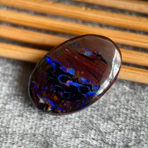 Peut inclure: Pierre précieuse ovale polie, avec une base brun foncé et des motifs bleus et oranges vifs. La surface de la pierre reflète la lumière, révélant des structures internes complexes. Probablement une opale.