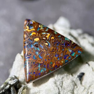 May include: A triangular, polished opal gemstone with a complex pattern of brown, yellow, blue, and orange hues. The stone's surface displays a network of intricate lines and shapes, creating a unique, natural design.