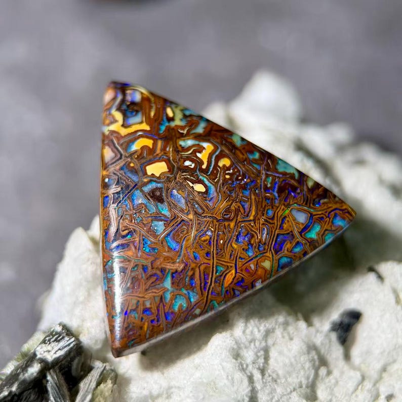 May include: A triangular, polished opal gemstone with a complex pattern of brown, yellow, blue, and orange hues. The stone's surface displays a network of intricate lines and shapes, creating a unique, natural design.