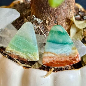 May include: Two triangular-shaped gemstones with layered colors of green, blue, and red-brown. The stones are set against a backdrop of a white pot and plant matter. The gemstones have a polished, smooth surface.