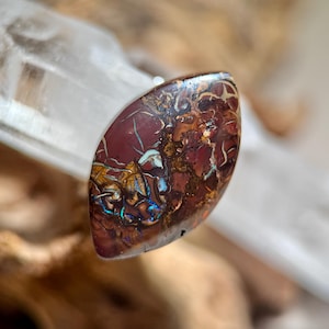 Peut inclure: Une pierre précieuse ovale polie, présentant une palette de couleurs riche et terreuse. La pierre a une base bordeaux foncé, entrelacée de veines blanches, dorées et des reflets bleus irisés. La surface est lisse et réfléchissante.