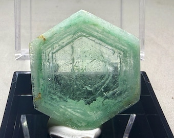 Puede incluir: Cristal verde translúcido hexagonal con estructuras internas visibles. El cristal está montado sobre una pequeña piedra blanca y sobre un fondo negro. Los bordes del cristal tienen un tono verde ligeramente más oscuro.