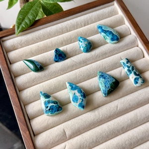 Puede incluir: Una colección de piedras preciosas pulidas de varias formas y tamaños, exhibidas en una bandeja de madera con un forro de terciopelo color crema. Las piedras presentan una mezcla de patrones azules, turquesas y blancos, algunos con apariencia moteada o veteada. Las piedras están bien dispuestas.